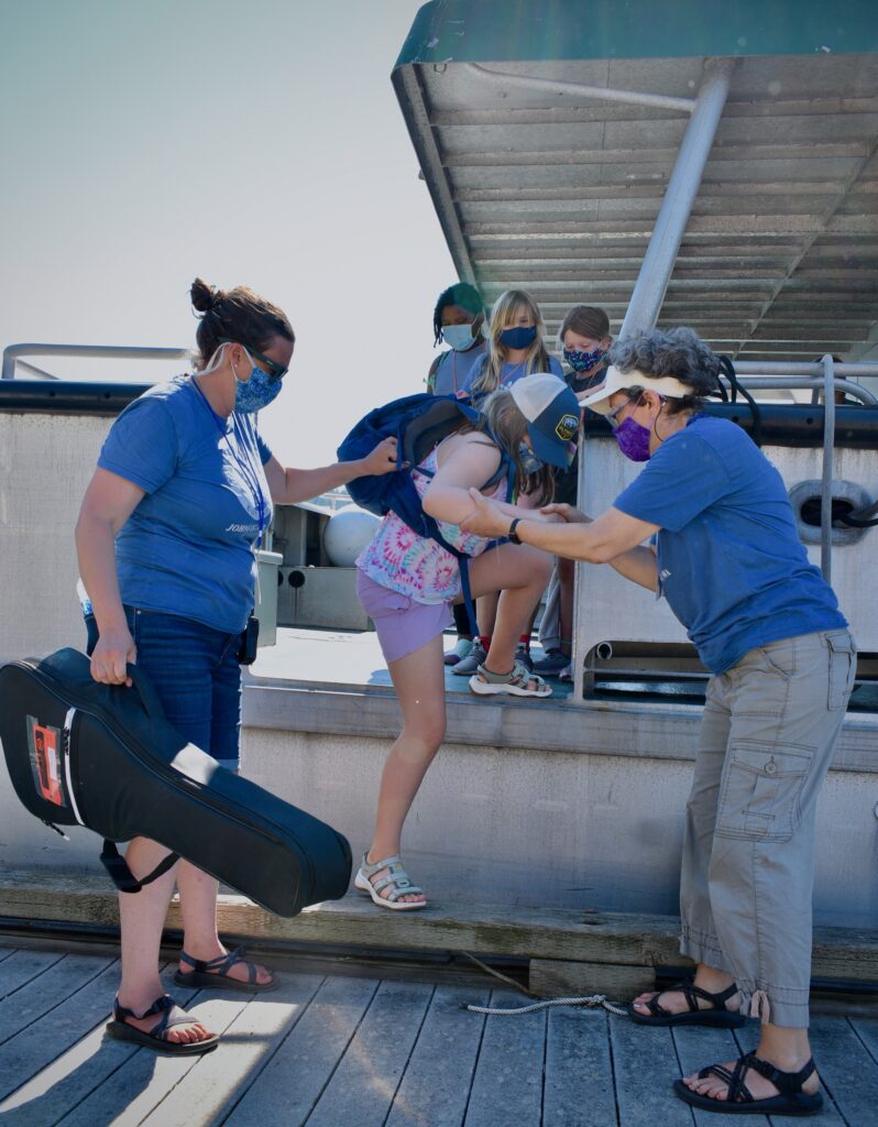 Two women wearing masks help a masked camper disembark from a boat onto a dock