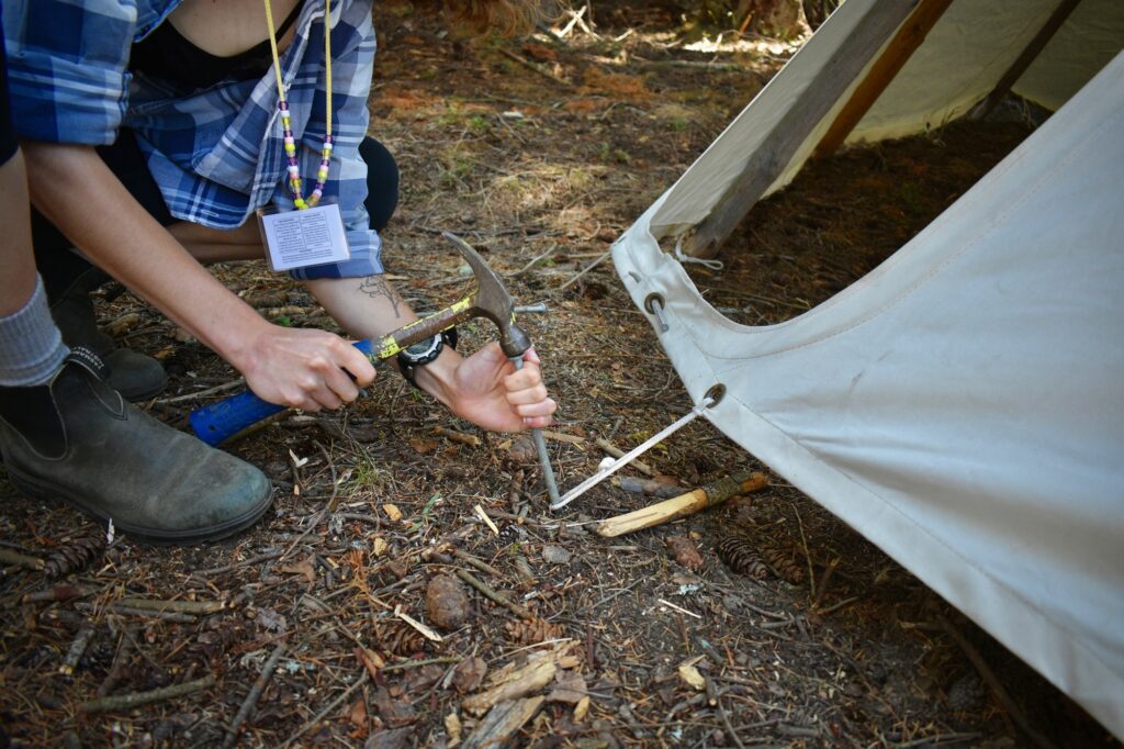 Close up of a staff member's hands nailing a metal stake into a rope loop at the base of a canvas tipi