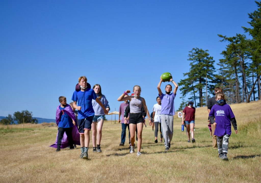 Campers dressed in purple walk down a field together as one raises a watermelon above their head in triumph