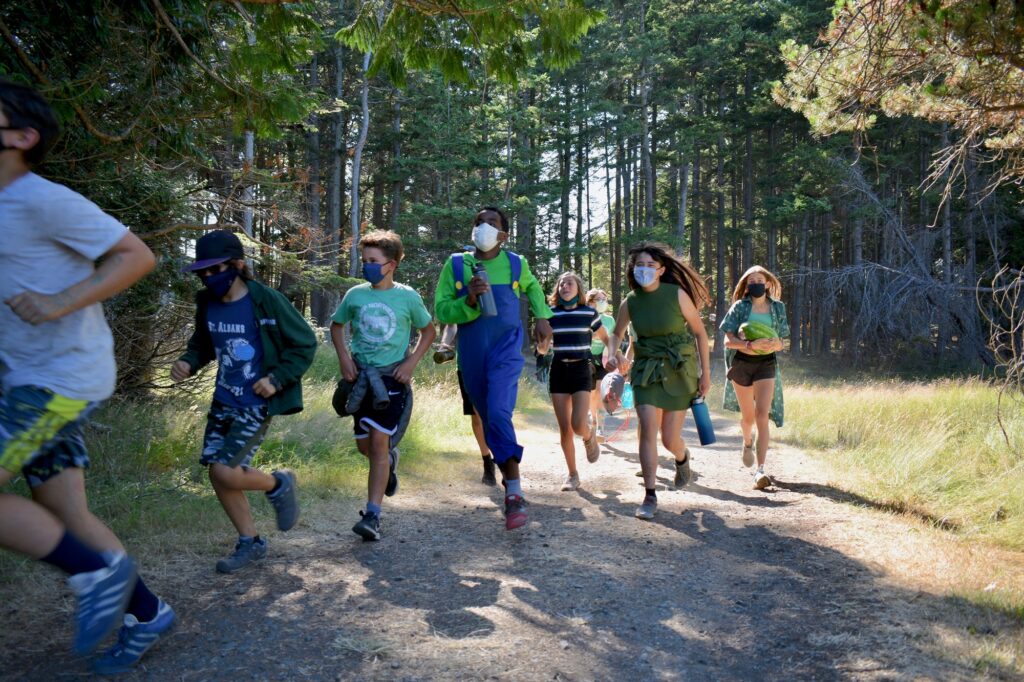 Campers dressed in green and wearing masks run down a road together while one carries a watermelon