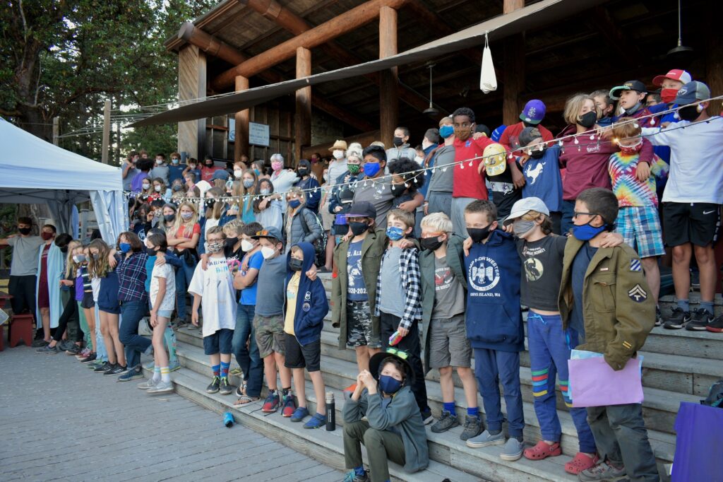 A large group of campers wearing masks stands together with their arms around each other on the steps of the Lodge