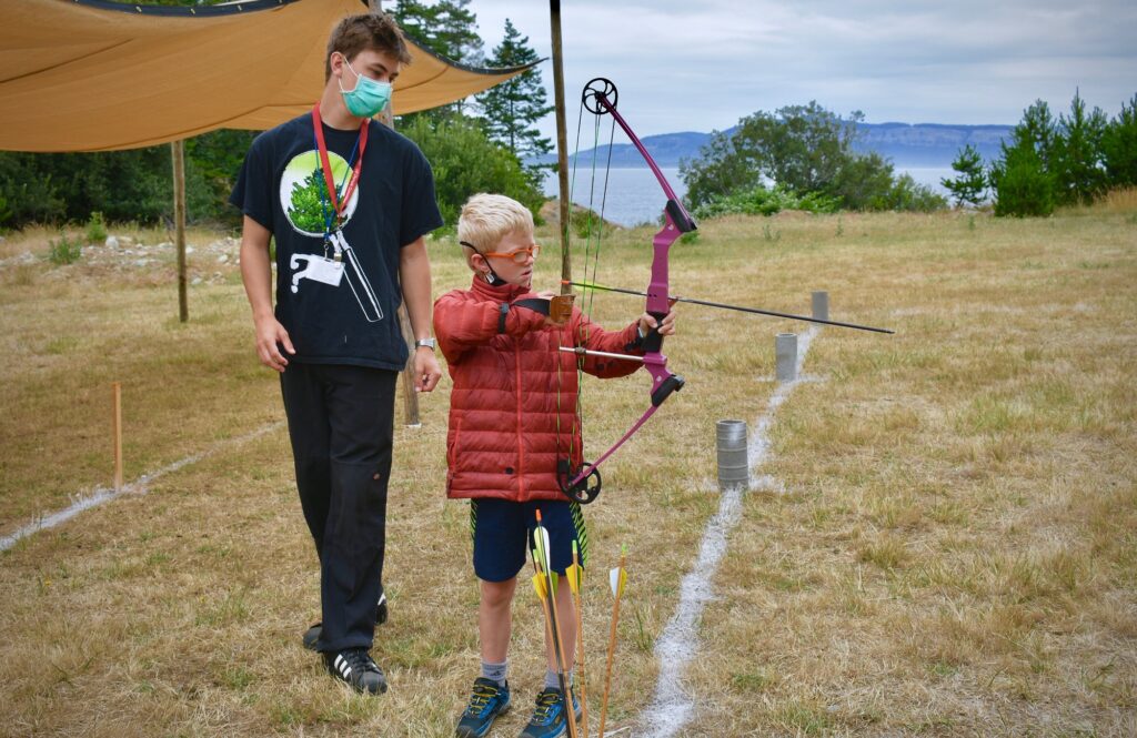 Staff member supervises a young camper drawing a bow at the archery range