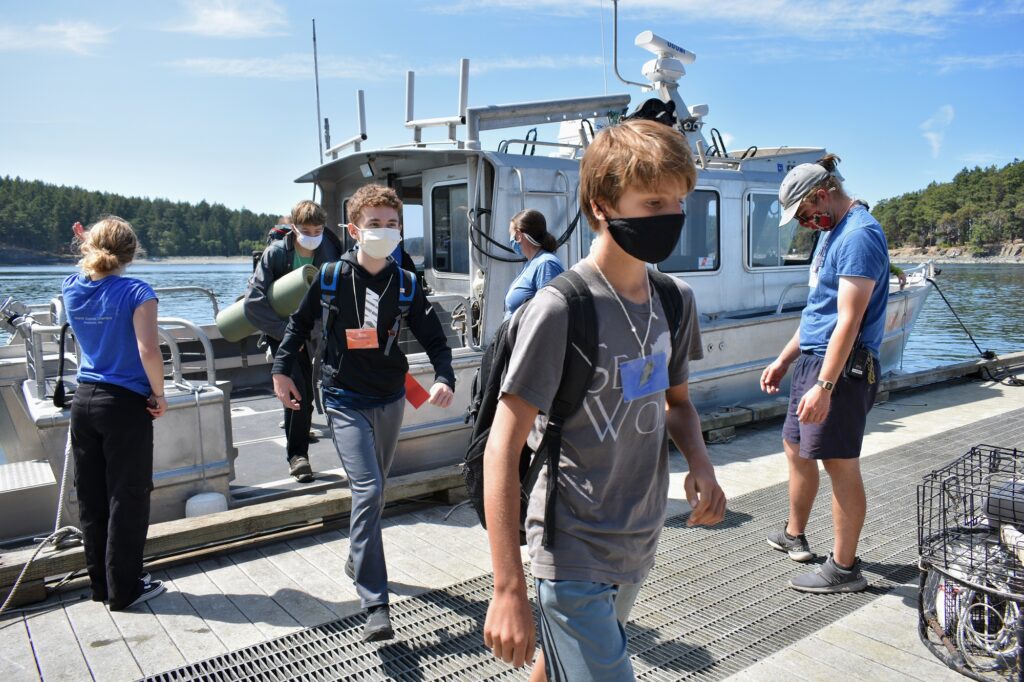 Three masked campers carrying gear step off a boat onto a dock as three staff members look on