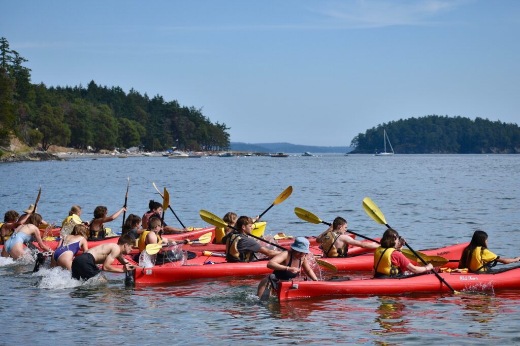 Campers in red kayaks push off from the beach at the start of the kayak race