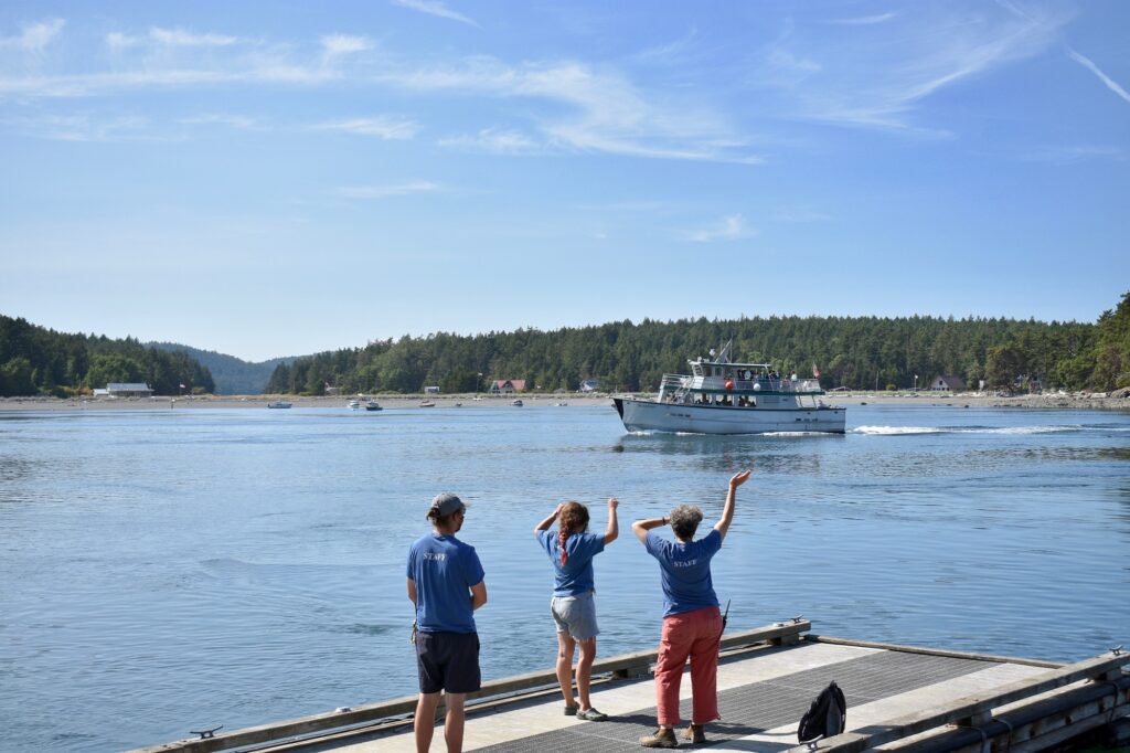 Three staff members stand on a dock and wave at a large boat in the distance
