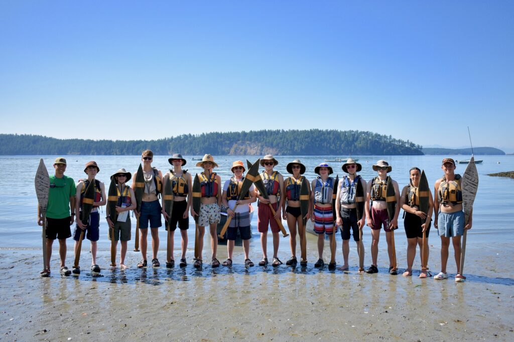 A line of people in hats and lifejackets pose holding canoe paddles on a beach