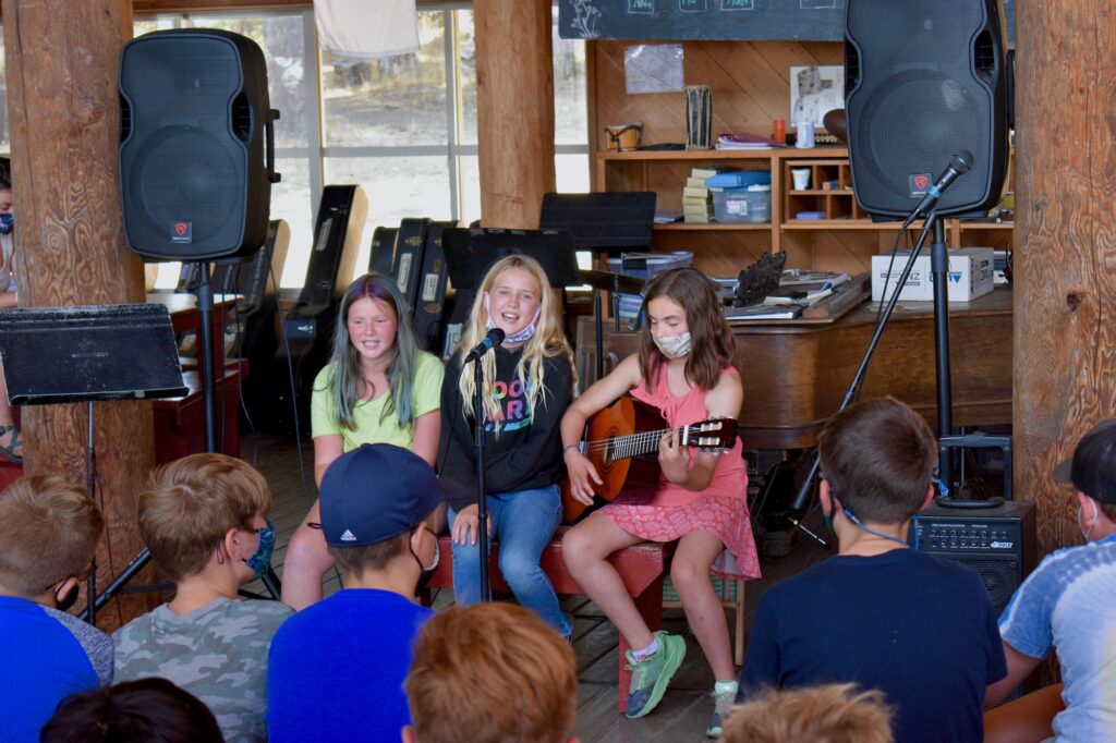 Three campers sing into a microphone while one plays guitar in front of an audience