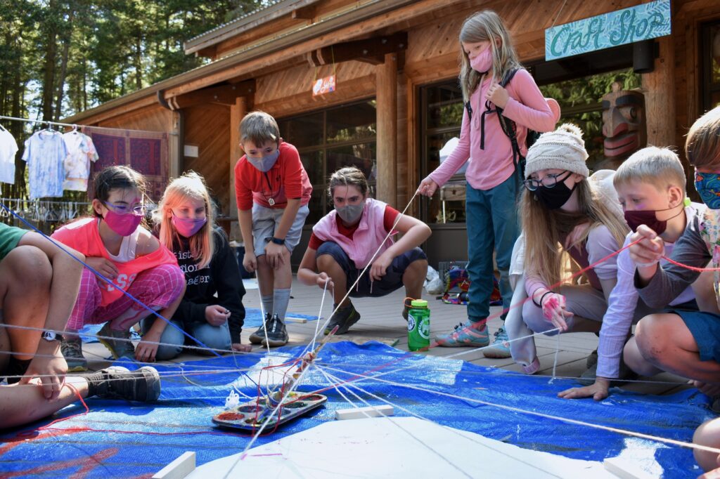 Campers dressed in pink and wearing masks hold pieces of yarn that connect in a spiderweb above a blue tarp