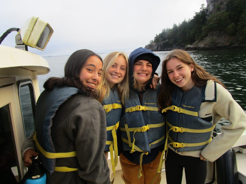 Four campers wearing life jackets pose and smile on the back deck of a boat