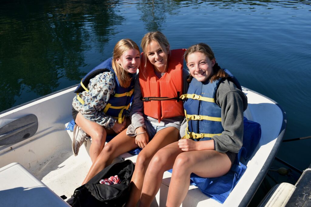 Three campers wearing life jackets smile as they sit together in the bow of a small boat