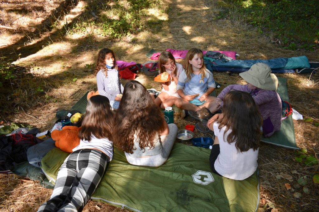 Several young campers sit on sleeping bags and eat food from cook kits
