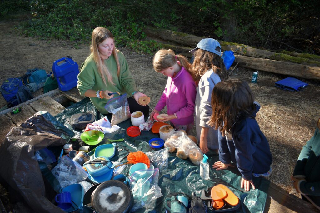 A staff member prepares breakfast items for three young campers while seated at a picnic table full of food and gear