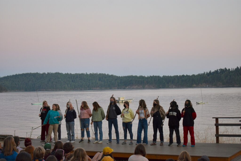 A group of masked campers stands in a line on a stage in front of water