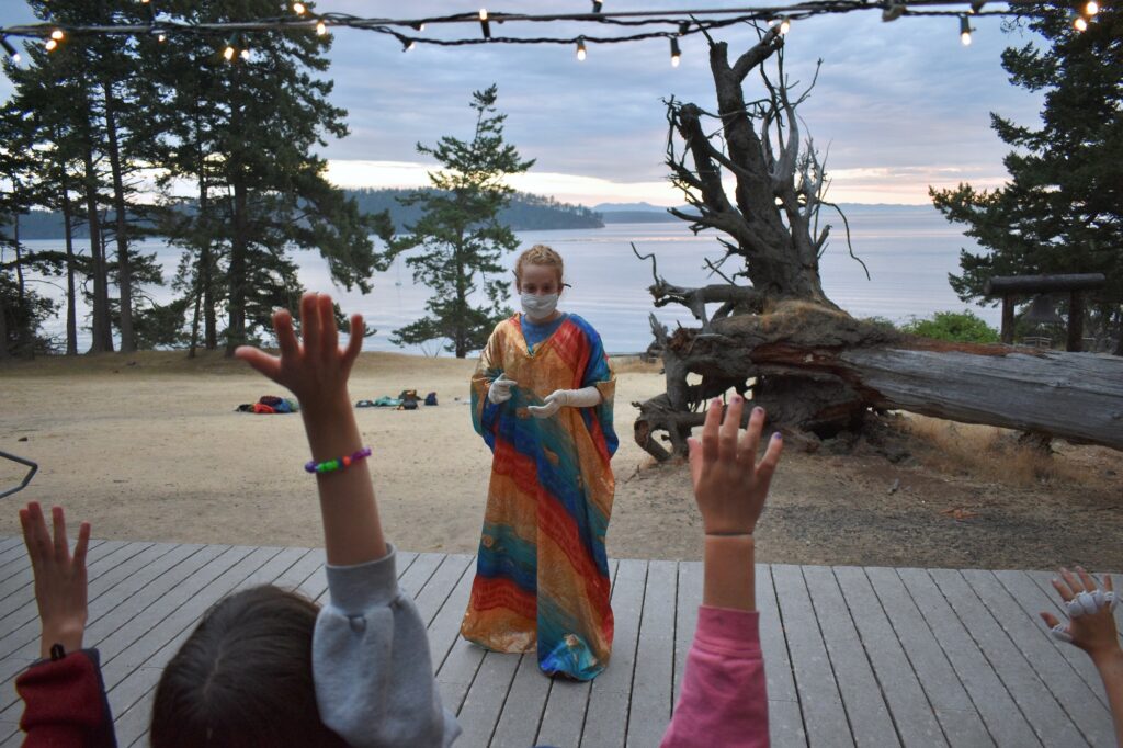 A masked camper wearing a colorful robe and long gloves stands onstage under a strand of fairy lights while other campers raise their hands in the foreground