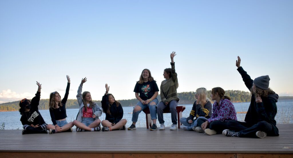 A group of campers raises their hands and laughs while seated on an outdoor stage