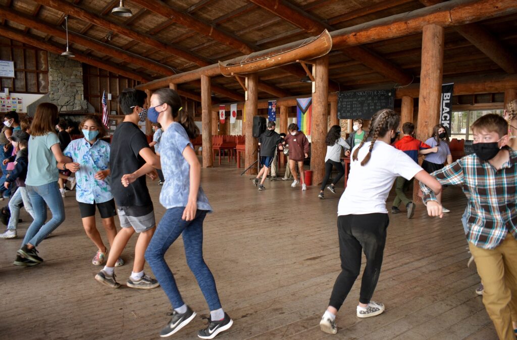 Campers wearing masks link arms and dance in pairs inside the Lodge