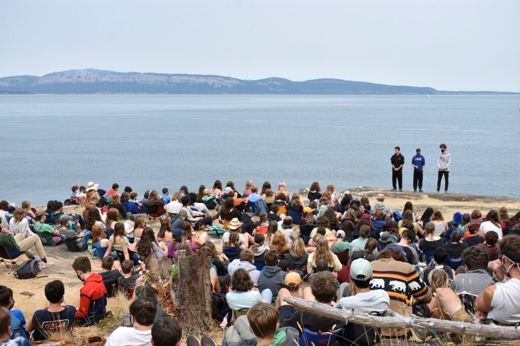 A trio of campers, with the ocean behind them, faces a large group of people sitting on the ground.