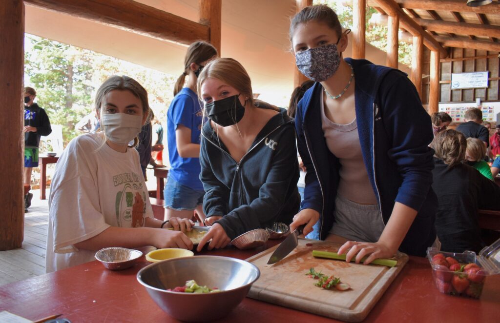 Three campers wearing masks cut up vegetables on a cutting board atop a red table