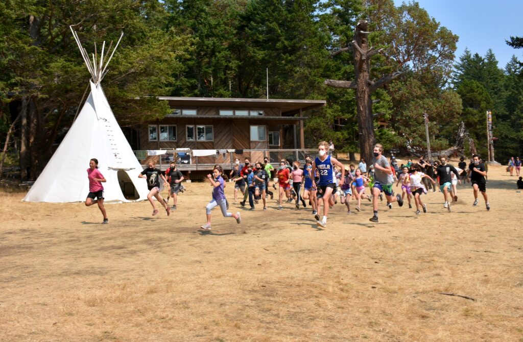 A large group of campers runs past a tipi and a brown building in the background