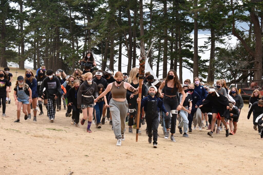 A large group of masked campers wearing black runs toward the camera