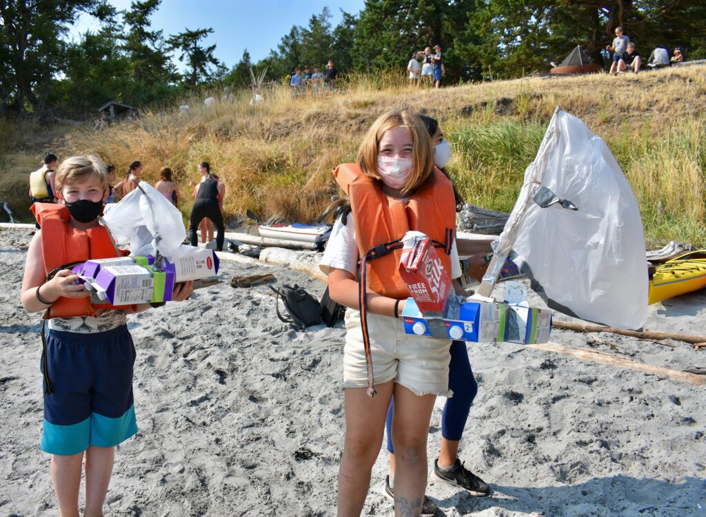 Campers wearing masks and orange life jackets smile as they hold up boats made out of milk cartons, sticks, and plastic bags