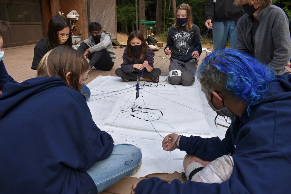 A group of masked campers gathers around a large sheet of paper and leads a pen held by multiple strings to make a drawing on the paper