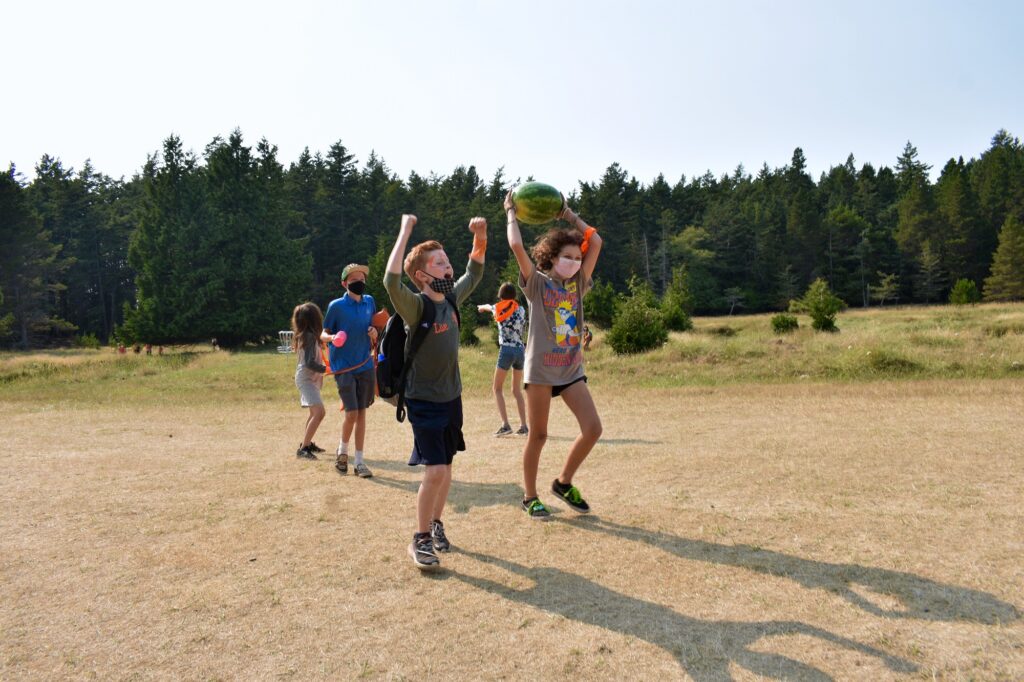 One camper raises their arms and yells victoriously while another holds a watermelon in triumph above their head